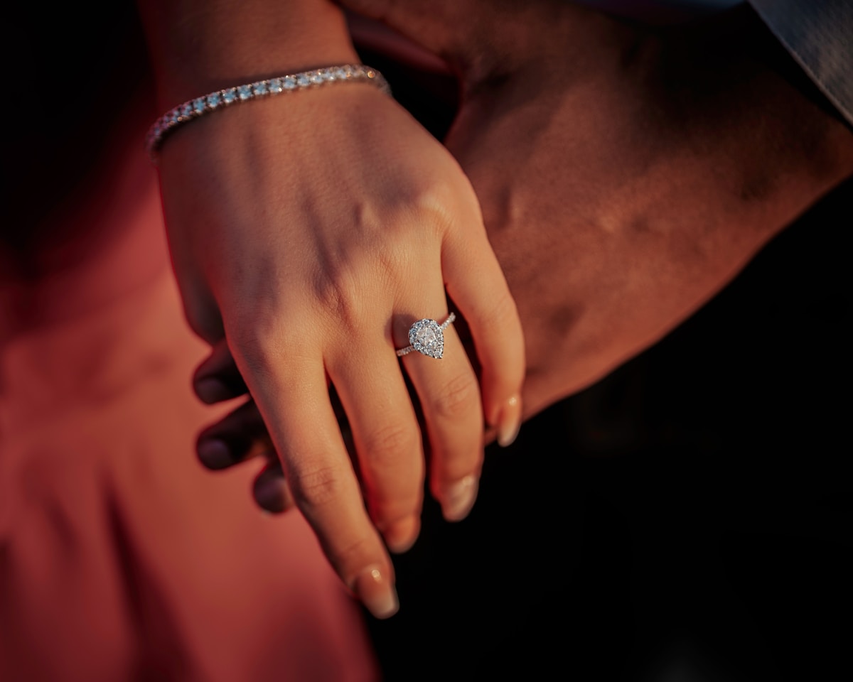 A couple holding hands, the woman's halo engagement ring visible in golden light