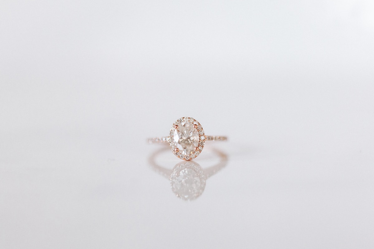 A rose gold oval halo engagement ring reflected on a white surface