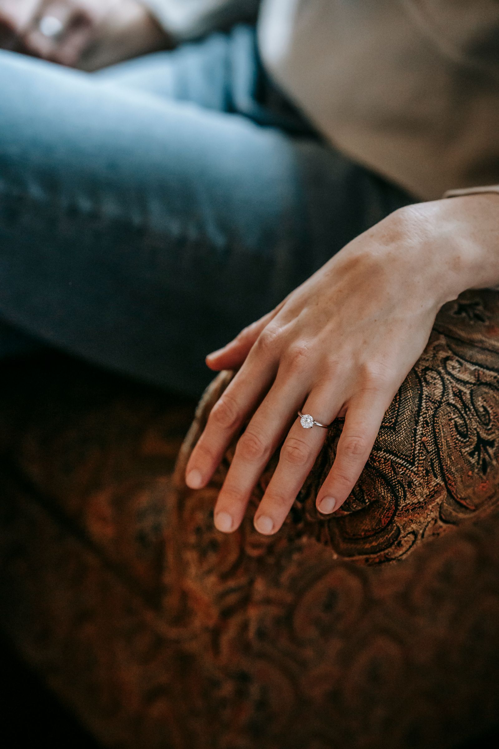 A woman's hand with a solitaire engagement ring resting casually on a patterned armchair